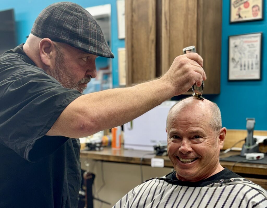 Barber in plaid cap giving a smiling client a close haircut at Venice Barbershop, classic blue wall, vintage decor, community vibe