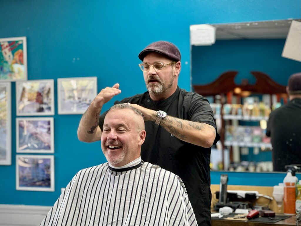 Barber giving a haircut to a smiling middle-aged man at Venice Barbershop, blue wall, framed artwork, and friendly vibe