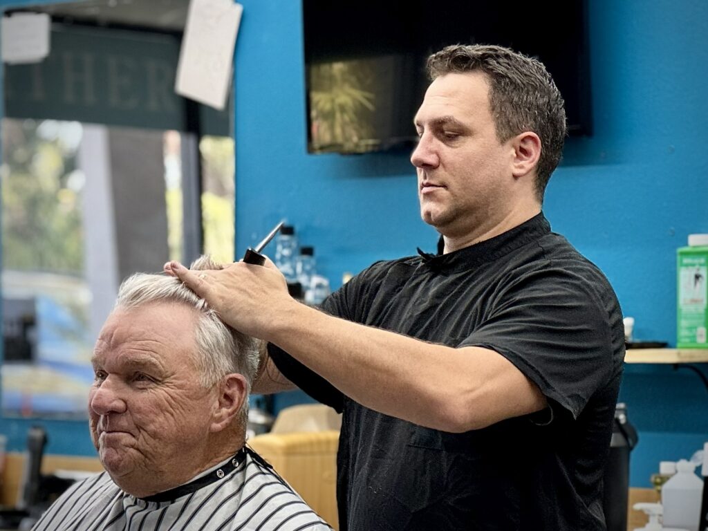 Barber giving a haircut to an older man at Venice Barbershop, blue wall background, professional and friendly atmosphere
