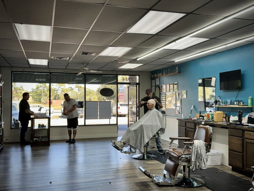 Wide view of Venice Barbershop interior, barber giving a haircut, customers chatting, blue wall, large windows, classic community barbershop