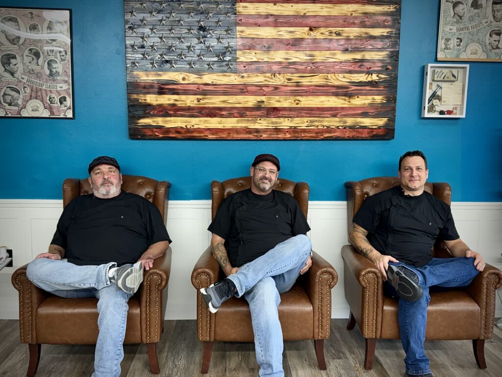 Three barbers sitting in brown leather chairs at Venice Barbershop, blue wall, large wooden American flag, classic barbershop posters