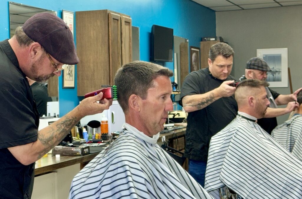 Three barbers giving haircuts to clients at Venice Barbershop, blue walls, classic striped capes, friendly and professional atmosphere