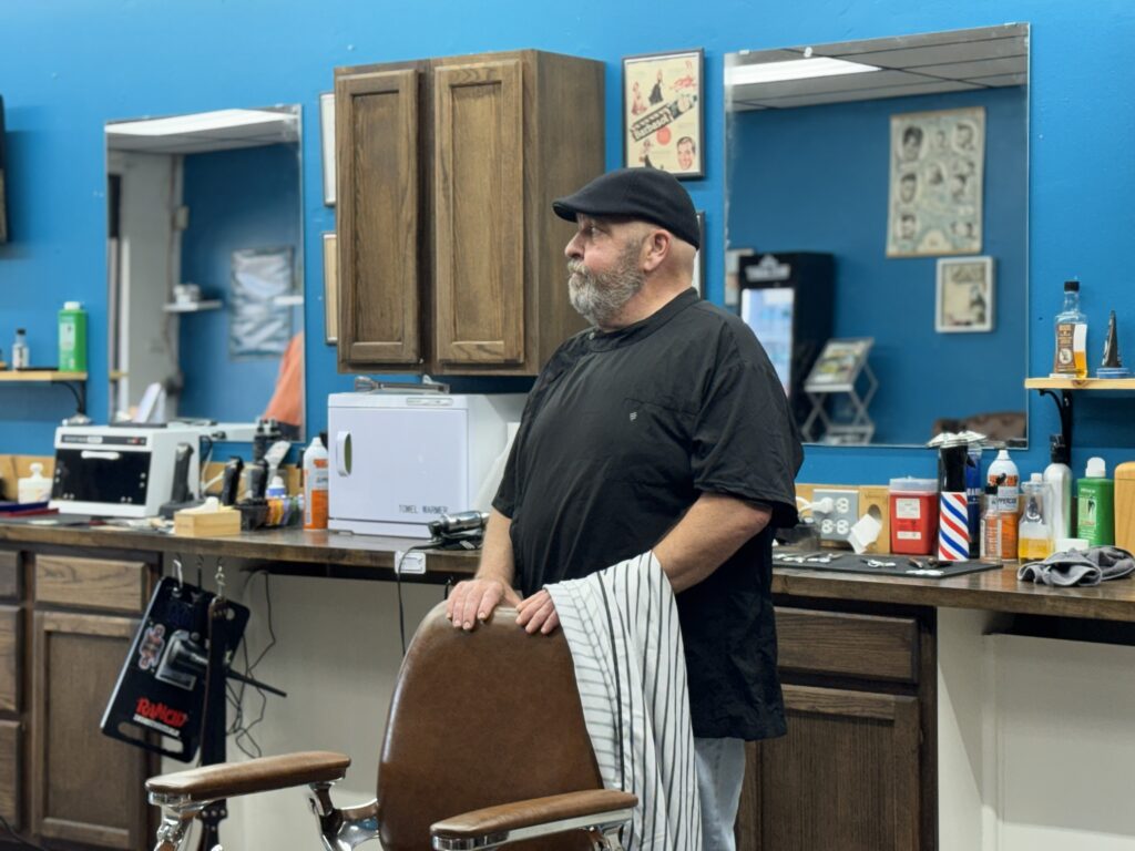 Barber in black shirt and cap standing behind a barber chair at Venice Barbershop, blue wall, classic barber tools on the counter