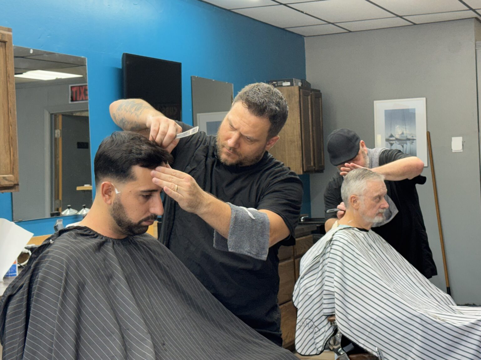 Barbers giving haircuts to two men, one younger and one older, at Venice Barbershop in Florida, classic shop with blue walls and wooden cabinets